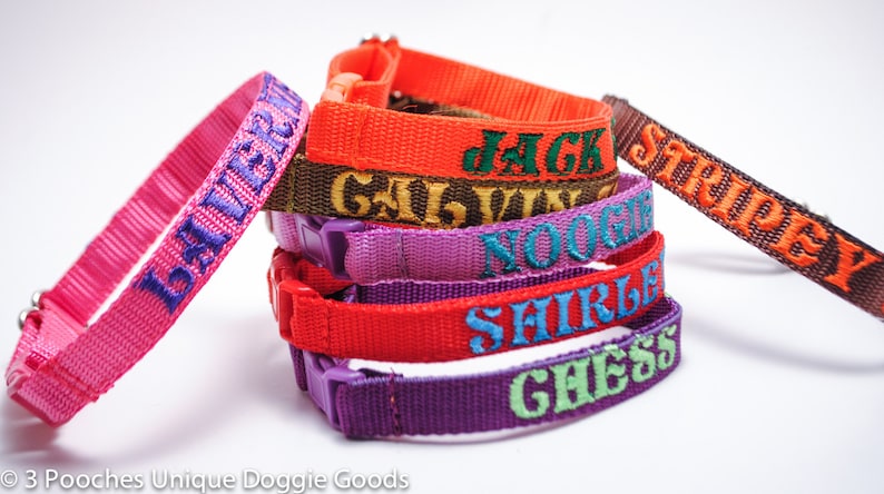 May include: A selection of colourful dog collars, each embroidered with a different name. The collars are in shades of pink, orange, purple, and red, with contrasting embroidery. They have plastic buckles and are made of a woven material.