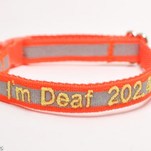 May include: An orange pet collar with reflective gray stripes and yellow embroidered text that reads "I'm Deaf 20245". The collar has a plastic buckle and a metal loop.