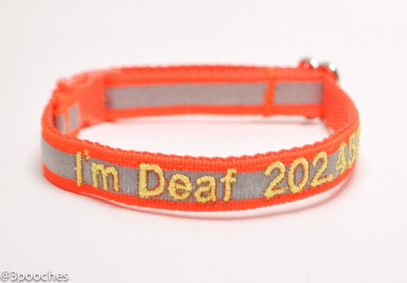 May include: An orange pet collar with reflective gray stripes and yellow embroidered text that reads "I'm Deaf 20245". The collar has a plastic buckle and a metal loop.