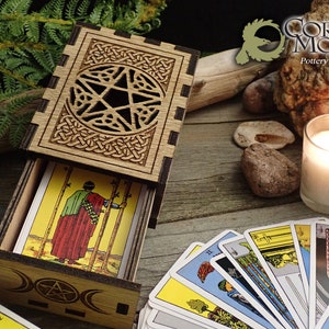 May include: Wooden box with a pentagram design and a sliding drawer. The drawer is open and contains a deck of tarot cards. The box is on a wooden surface with a candle and rocks.