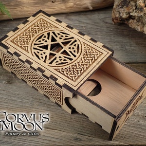 May include: A wooden box with a sliding drawer, intricately carved with a Celtic knot design and a pentagram. The box is made of light wood with dark brown accents.