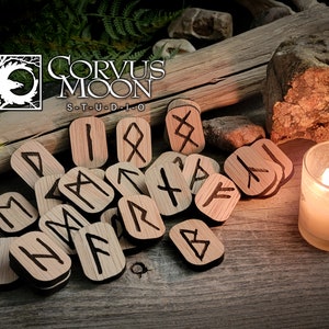 May include: Wooden rune stones with black etched symbols are scattered on a wooden surface. The Corvus Moon Studio logo is in the upper left. A lit candle in a glass holder is in the lower right. The runes are for divination.