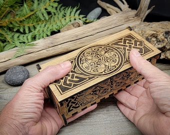 May include: A wooden box with an intricate Celtic knot design carved into the lid. The box is made of light brown wood and has a dark brown stain. The box is being held in a person's hands.