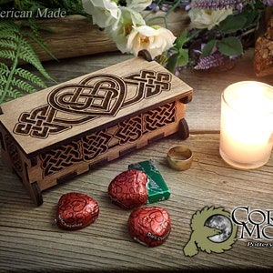 May include: A wooden box with a Celtic knot design featuring a heart. The box is closed and sitting on a wooden surface with a candle, a ring, and chocolate hearts. The text "Corvus Moon Pottery & Gifts" is visible in the lower right corner.
