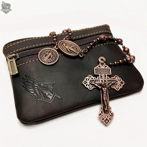 Peut inclure: Une pochette en cuir marron foncé avec une fermeture éclair en bronze et un chapelet assorti. Le chapelet comprend un crucifix et plusieurs médaillons. Un écusson avec des ailes et une épée est estampé sur la pochette.