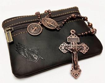 May include: A dark brown leather pouch with a bronze zip and a matching rosary. The rosary features a crucifix and several medallions. A shield with wings and a sword is embossed on the pouch.