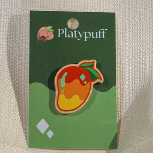 May include: A mango-shaped pin with red, orange, and yellow colors, and green leaves. The pin is on a green card with the text "Platypuff" and a small illustration of a fruit. The background is a textured, off-white fabric.
