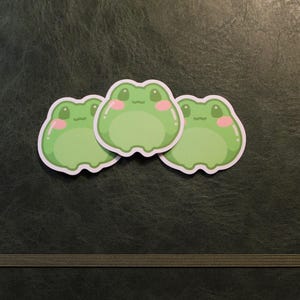 Può includere: Tre adesivi a forma di rana verde con guance rosa e contorni bianchi sono disposti su una superficie strutturata verde scuro. Gli adesivi hanno un design a cartone animato, con un'espressione semplice e amichevole.