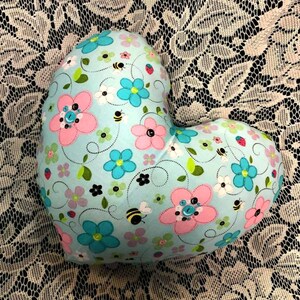 Puede incluir: Una almohada en forma de coraz&oacute;n con un fondo azul claro y un patr&oacute;n floral. Las flores son rosas, azules y blancas con hojas verdes y peque&ntilde;as abejas negras. La almohada est&aacute; decorada con botones.