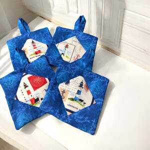 May include: Four blue fabric pot holders with a white background and a lighthouse design. Each pot holder has a different lighthouse design with text that says "Beach Island", "The Sea", "Lighthouse Cove", and "Sweet Escape".