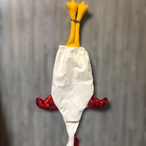 Puede incluir: Un pa&ntilde;o de cocina de tela blanca con forma de pollo, con alas rojas, un pico amarillo y patas amarillas. El pa&ntilde;o est&aacute; colgado boca abajo.