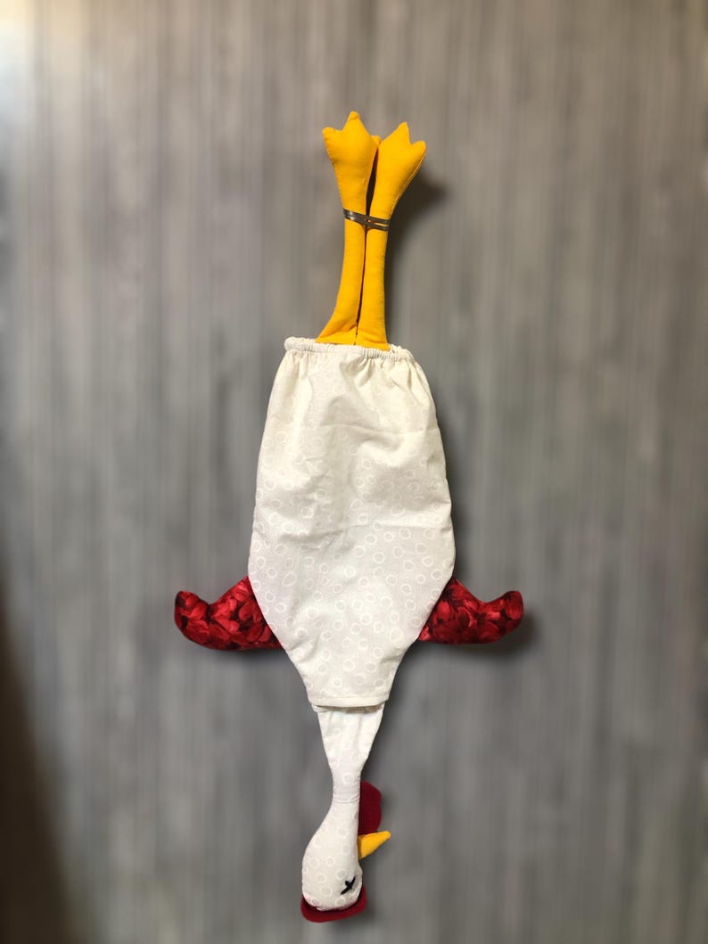 Puede incluir: Un pa&ntilde;o de cocina de tela blanca con forma de pollo, con alas rojas, un pico amarillo y patas amarillas. El pa&ntilde;o est&aacute; colgado boca abajo.
