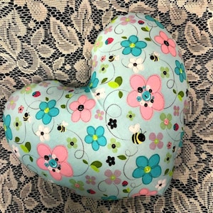 Puede incluir: Una almohada con forma de coraz&oacute;n con un fondo azul claro y un patr&oacute;n floral. El patr&oacute;n presenta flores rosas, azules y blancas, abejas y hojas.