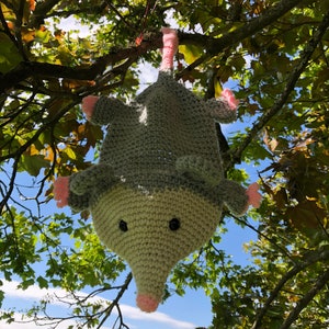 May include: A crocheted gray and pink opossum hanging from a tree branch. The opossum has black eyes and a long, pink nose.