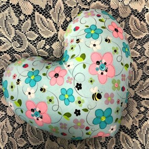 Puede incluir: Una almohada con forma de coraz&oacute;n con un fondo azul claro y un patr&oacute;n floral. El patr&oacute;n presenta flores rosas, azules y blancas, abejas y hojas.