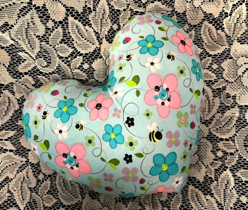 Puede incluir: Una almohada con forma de coraz&oacute;n con un fondo azul claro y un patr&oacute;n floral. El patr&oacute;n presenta flores rosas, azules y blancas, abejas y hojas.