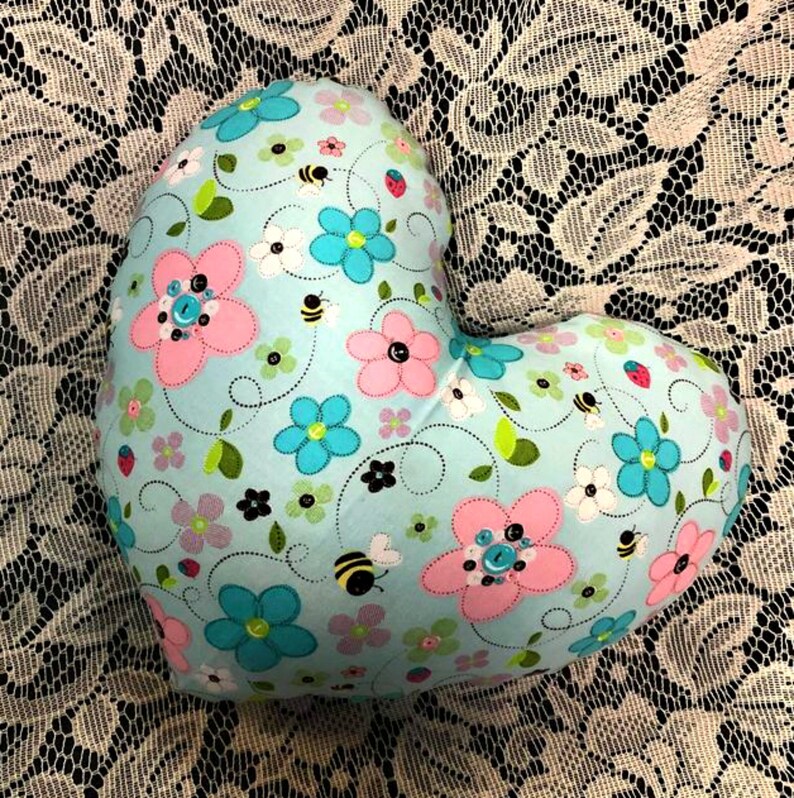 Puede incluir: Una almohada en forma de coraz&oacute;n con un fondo azul claro y un patr&oacute;n floral. Las flores son rosas, azules y blancas con hojas verdes y peque&ntilde;as abejas negras. La almohada est&aacute; decorada con botones.