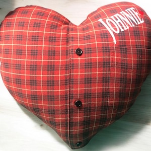 Puede incluir: Almohada en forma de coraz&oacute;n de cuadros rojos y negros con el nombre "JOHNNIE" bordado en blanco en el lado derecho. La almohada tiene dos botones negros.