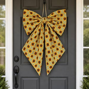 May include: A large decorative bow made of fabric featuring a repeating pattern of yellow sunflowers with brown centers set against a small green and white gingham check background. The bow has two wide top loops and two long, pointed hanging tails.