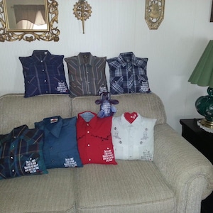 May include: A collection of seven square throw pillows crafted from repurposed button-down shirts in various colors and patterns, including plaid, solid red, and white. Each pillow features a collar, buttons, and a printed memorial message.