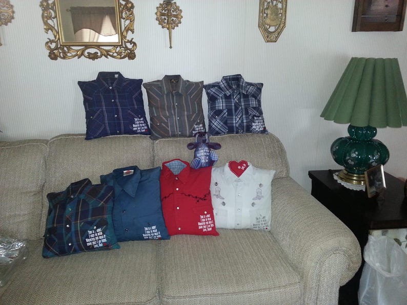 May include: A collection of seven square throw pillows crafted from repurposed button-down shirts in various colors and patterns, including plaid, solid red, and white. Each pillow features a collar, buttons, and a printed memorial message.