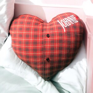 Puede incluir: Almohada en forma de coraz&oacute;n de cuadros rojos y negros con la palabra "JOHNNIE" bordada en blanco en la parte delantera.
