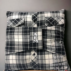Pode incluir: Uma almofada de xadrez preto e branco com um bolso de cada lado e um fecho de bot&atilde;o. A almofada tem as iniciais "AEM" bordadas na frente e o texto "This is a shirt I used to wear. Whenever you hold it know that I am there. Love, Pawdaw" impresso na parte inferior.