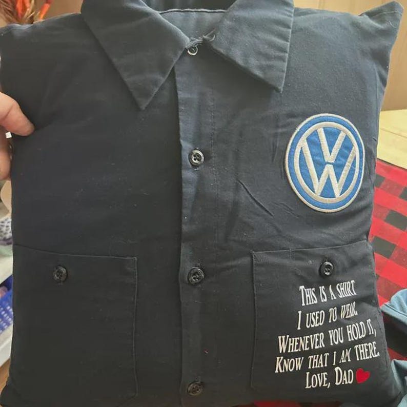 May include: A dark navy blue square pillow constructed from a repurposed button-down shirt. It features a collar, a central button placket, two front pockets, a circular Volkswagen logo patch, and white text that reads: This is a shirt I used to wear. Whenever you hold it, know that I am there. Love, Dad.