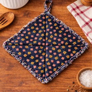 May include: A navy blue, square-shaped quilted potholder with a floral pattern of yellow sunflowers and red squares. The edges are trimmed with a frayed, multi-colored fabric. A loop is attached for hanging. Kitchen decor.