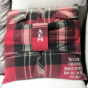 Pode incluir: Uma almofada de xadrez vermelho e preto com um bolso vermelho na frente. O bolso tem as letras "AEM" bordadas em branco. A almofada tem uma mensagem na parte inferior que diz "This is a shirt I used to wear. Whenever you hold it know that I am there. Love, Dad."
