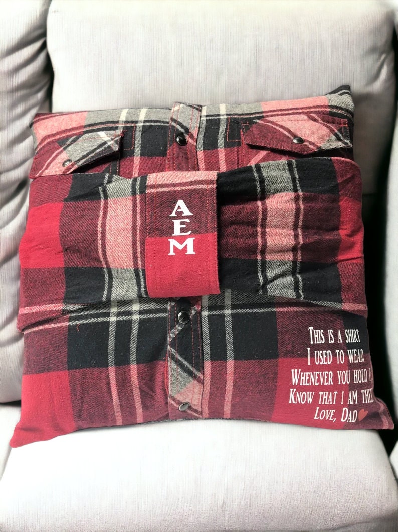 Pode incluir: Uma almofada de xadrez vermelho e preto com um bolso vermelho na frente. O bolso tem as letras "AEM" bordadas em branco. A almofada tem uma mensagem na parte inferior que diz "This is a shirt I used to wear. Whenever you hold it know that I am there. Love, Dad."