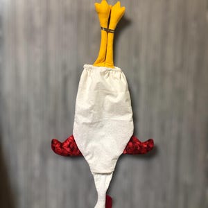 Puede incluir: Un pa&ntilde;o de cocina de tela blanca con forma de pollo, con alas rojas, un pico amarillo y patas amarillas. El pa&ntilde;o est&aacute; colgado boca abajo.