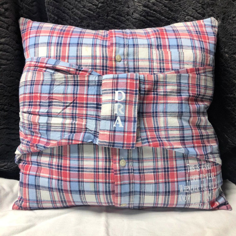 Pode incluir: Uma almofada de xadrez vermelho, azul e branco com uma manga de camisa dobrada no topo. A manga tem o nome "DRA" bordado. A almofada tem o texto "This is a shirt I used to wear. Whenever you hold it know I am with you. Love, Dad" bordado na parte inferior.