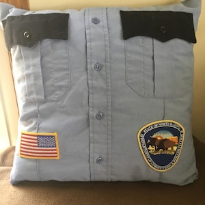 May include: A light blue pillow made from a button-down shirt, featuring black pocket flaps and buttons. An American flag patch and a North Dakota Department of Corrections & Rehabilitation patch are sewn on the pillow.