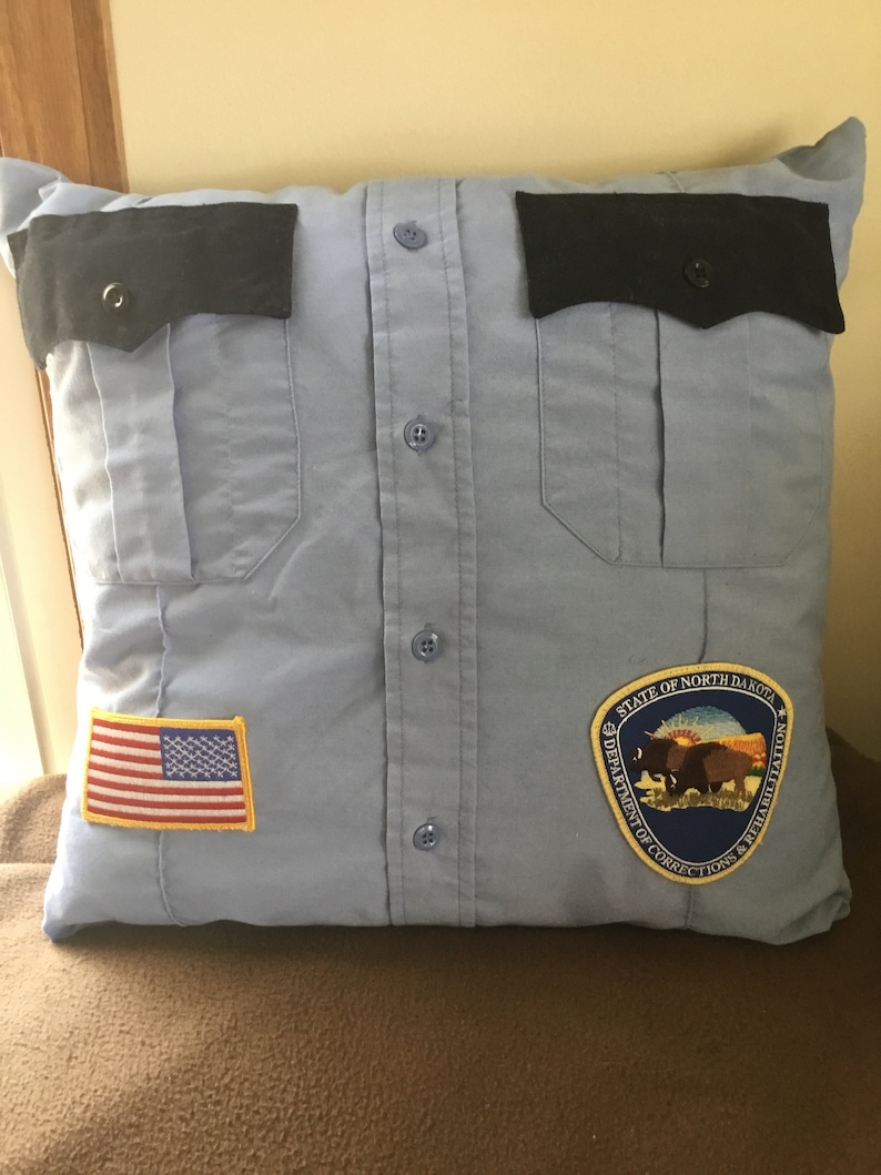 May include: A light blue pillow made from a button-down shirt, featuring black pocket flaps and buttons. An American flag patch and a North Dakota Department of Corrections & Rehabilitation patch are sewn on the pillow.