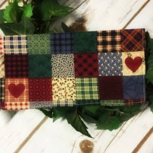 May include: A patchwork fabric wallet with a red heart design. The wallet is made of various fabrics in shades of red, green, blue, and brown.