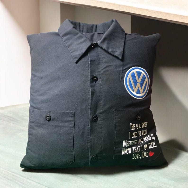 May include: A black square pillow shaped like a button-down shirt with a collar, a central button placket, and two front pockets. A blue and white Volkswagen logo is on the upper right, and white text reads: This is a shirt I used to wear. Whenever you hold it, know that I am there. Love, Dad with a red heart.