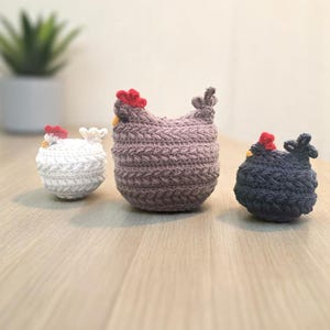 May include: Three crocheted chicken figurines in white, purple, and navy blue. Each chicken has a red comb and a yellow beak. The figurines are sitting on a light brown wooden surface.