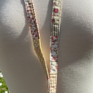May include: A floral lanyard with a variety of patterns. One side features a cream background with pink flowers, while the other has a cream background with yellow polka dots. The lanyard is designed to be worn around the neck.