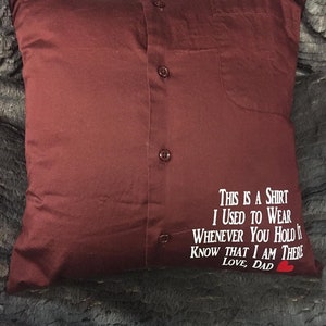 May include: A burgundy pillow made from a button-down shirt. The shirt's buttons and pocket are visible. White text reads "THIS IS A SHIRT I USED TO WEAR WHENEVER YOU HOLD IT KNOW THAT I AM THERE LOVE, DAD" with a red heart.
