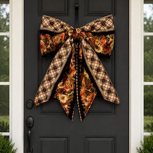 May include: A large decorative bow hanging on a black door. The bow features a black fabric base with a vibrant autumn-themed print of pumpkins, sunflowers, and foliage, bordered by a classic brown, black, and white plaid pattern trim.