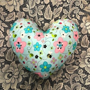 Puede incluir: Una almohada con forma de coraz&oacute;n con un fondo azul claro y un patr&oacute;n floral con flores rosas, azules y verdes, abejas y espirales.
