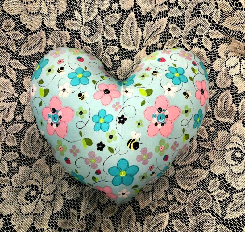 Puede incluir: Una almohada con forma de coraz&oacute;n con un fondo azul claro y un patr&oacute;n floral con flores rosas, azules y verdes, abejas y espirales.