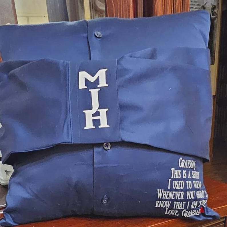 Pode incluir: Uma almofada azul com um design de camisa bordada em branco. A camisa tem um bolso com as iniciais "MJH" bordadas. A almofada tem uma mensagem que diz "Grayson, esta &eacute; uma camisa que eu costumava usar, sempre que voc&ecirc; a segurar, saiba que estou l&aacute;. Com amor, Av&ocirc;."