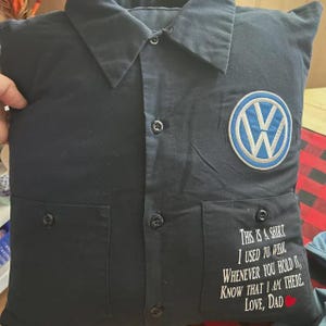 May include: A dark navy blue square pillow constructed from a repurposed button-down shirt. It features a collar, a central button placket, two front pockets, a circular Volkswagen logo patch, and white text that reads: This is a shirt I used to wear. Whenever you hold it, know that I am there. Love, Dad.