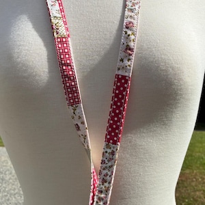 May include: A floral and polka dot patterned lanyard with a silver-colored clasp. The lanyard features a patchwork design with red, white, and floral fabric sections. The lanyard is displayed on a white mannequin.