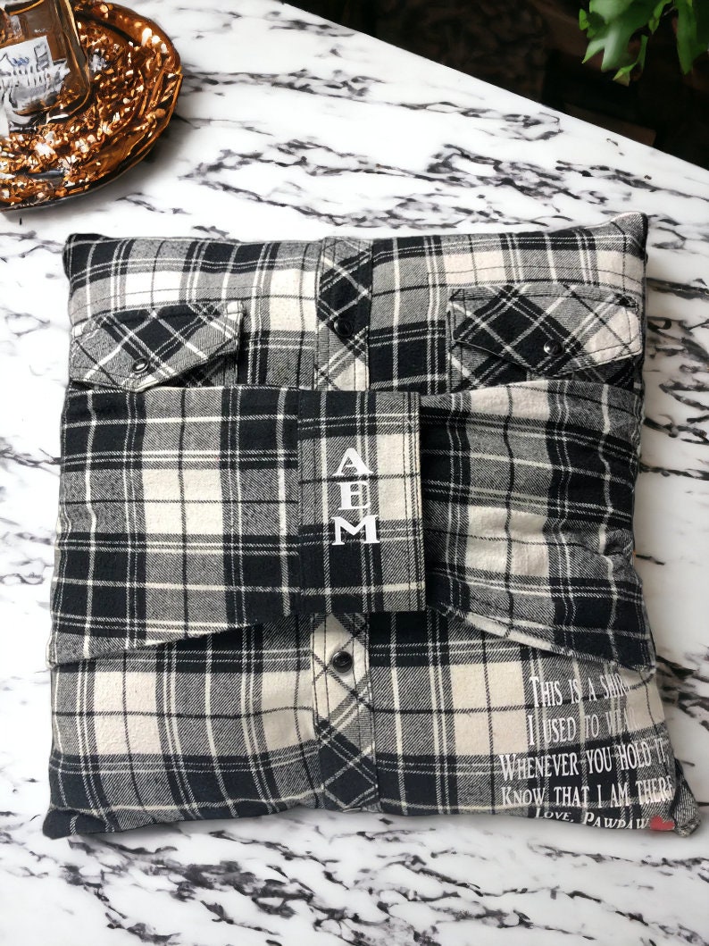 Pode incluir: Uma almofada de xadrez preto e branco com um bolso de cada lado. A almofada tem as iniciais "A B M" bordadas na frente. A parte de tr&aacute;s da almofada tem o texto "This is a shirt I used to wear. Whenever you hold it know that I am there. Love, Pawdaw" impresso nela.