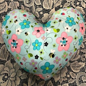 Puede incluir: Una almohada con forma de coraz&oacute;n con un fondo azul claro y un patr&oacute;n floral. El patr&oacute;n incluye flores rosas y azules, hojas verdes y abejas negras.