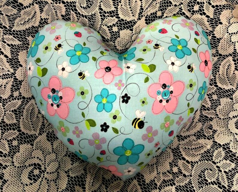 Puede incluir: Una almohada con forma de coraz&oacute;n con un fondo azul claro y un patr&oacute;n floral. El patr&oacute;n incluye flores rosas y azules, hojas verdes y abejas negras.