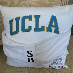 Pode incluir: Travesseiro branco com o texto "UCLA" em letras azuis e amarelas. O travesseiro tem um bolso na frente com as letras "SB" em preto. A parte de tr&aacute;s do travesseiro tem o texto "This is a shirt I used to wear whenever you hold it know that I am with you" em texto preto.